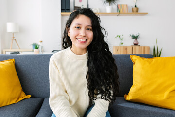 Portrait of cheerful young latin american woman smiling at camera sitting on sofa at home.
