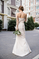 back view of young woman in white dress holding wedding bouquet behind back.