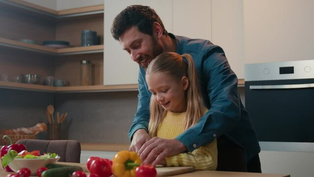 Adult father teaching Caucasian kid daughter cut fresh vegetable salad in kitchen cooking together. Parent dad learning small child girl cook healthy food help with cutting cucumber for vegan meal
