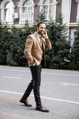 full length of cheerful man in beige formal wear standing with hand in pocket and looking at camera.