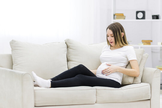 Pregnant woman touching her stomach on white sofa.