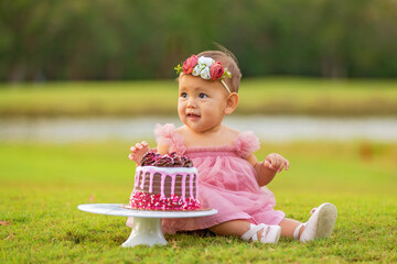 1 year old baby girl sitting behind birthday cake outdoors on green grass.
