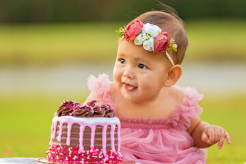 1 year old baby girl sitting behind birthday cake outdoors on green grass.