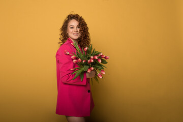 Portrait of a happy charming red-haired girl receiving flowers, a girl with pink tulips, happily smiling and laughing, on a yellow background in pink clothes.Holidays, beauty and spring concept