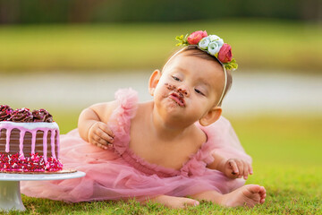 1 year old baby girl sitting behind birthday cake outdoors on green grass. Baby has cake funny expression and cake on face.