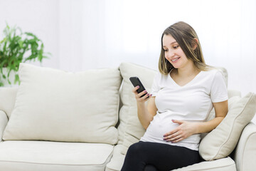 Pregnant woman sitting on white sofa looking at the phone.