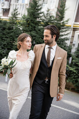 young bride in white dress holding wedding bouquet while walking with bearded groom on street.