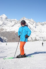 Fototapeta na wymiar Skier in blue jacket, black helmet and orange pantson the piste slope in winter with snow mountains in Alps, Europe