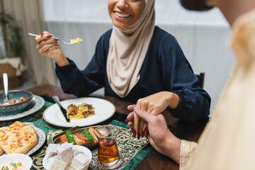Cropped view of muslim couple holding hands near food during iftar at home.