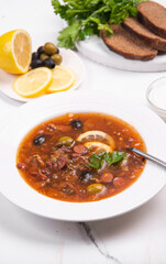 Solyanka in a white plate on a white table. Vegetable soup with olives for lunch.