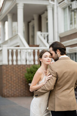 bearded groom in beige suit hugging charming bride in white wedding dress.