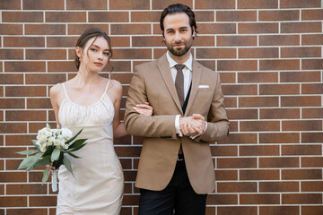 bride in wedding dress holding flowers near groom in suit while standing against brick wall.
