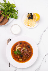 Solyanka in a white plate on a white table. Vegetable soup with olives for lunch.