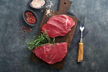 Raw beef steak. Marbled raw fresh Ribeye steak with rosemary, salt and pepper on cutting board on dark concrete background. Raw beef steak and spices for cooking.