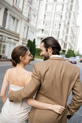 happy groom in beige suit hugging and looking at cheerful bride in white wedding dress.