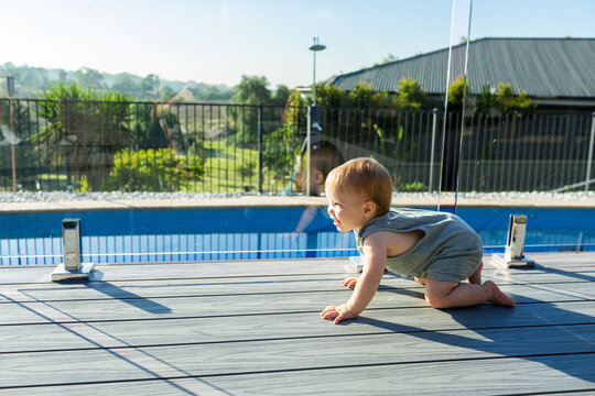 Happy baby crawling on deck beside glass pool fence in summer