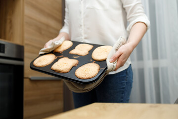 Woman holding hot tray with baked cupcakes, muffins, cookies in potholder, standing near oven in modern kitchen. Homemade cooking. Person with no face