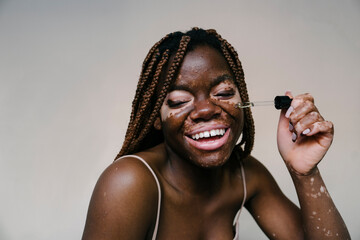 portrait of a pretty african woman with vitiligo applying an essence serum with a dropper while...