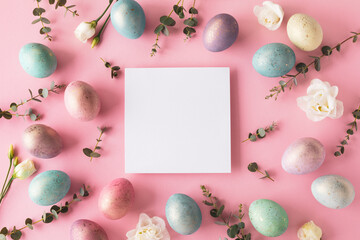 Easter background with easter eggs and flowers on pink table top view. White card for greeting and invitation. Flat lay.