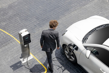 Fototapeta Aerial view of progressive businessman in black formal suit with his electric vehicle recharging battery at public car park charging station as vehicle powered by sustainable energy concept. obraz