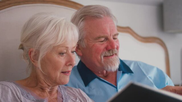 Close up of smiling retired senior couple in bed at home looking through photo album and reliving old memories - shot in slow motion