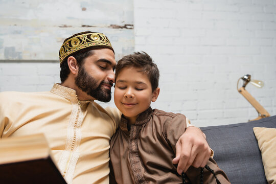 Smiling Muslim Man With Prayer Beads Hugging Son Near Book On Couch At Home.