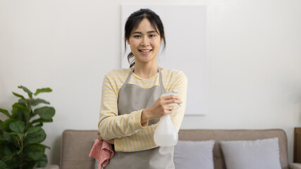 Obraz premium Asian housewife wearing apron holding rag and cleaning spray prepares for a big cleanup in the house, Housework, Daily routine, Big cleaning, Deep stain remover, Clean up on weekends.