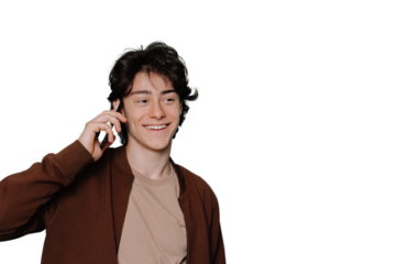 Cheerful young handsome guy in beige t-shirt and brown jacket talks by phone toothy smiles looks aside standing against blue studio backdrop. Brunette boy isolated on transparent background. Mockup.