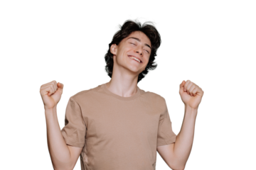 Happy Italian teen rises clenched fists in a winner expression stands against transparent background eyes closed. Amazed schoolboy celebrates win. Caucasian guy in beige t-shirt toothy smiles.