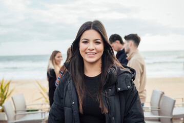 Portrait of a beautiful brunette girl smiling and looking at camera with a group of friends in a terrace bar on the beach