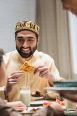 Smiling muslim father holding pita bread near blurred family during ramadan dinner.
