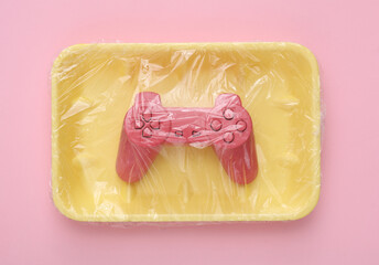 Video game gamepad in package wrapped with cellophane film on pink background. Creative idea. Minimal layout