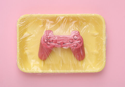 Video game gamepad in package wrapped with cellophane film on pink background. Creative idea. Minimal layout