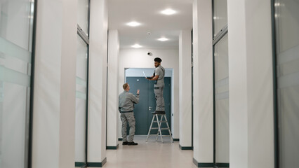 Two multi ethnic installers in uniform sets up security camera in office hallway using laptop. They talk, check CCTV cameras in computer program. Concept of surveillance system and privacy.