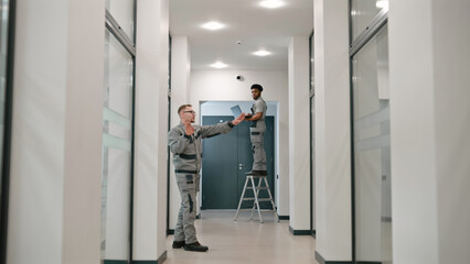 Two multi ethnic installers in uniform sets up security camera in office hallway using laptop. They talk, check CCTV cameras in computer program. Concept of surveillance system and privacy.