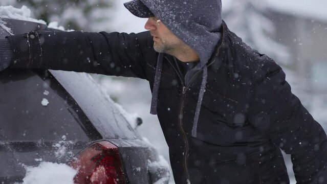 Man in hoodie cleaning car from snow with brush tool during heavy snowfall cyclone slow motion. Snowstorm in winter season. Vehicle care in blizzard