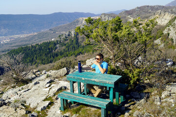 Fototapeta na wymiar A tourist drinks a drink from a thermos, sitting on a bench in a picturesque mountainous area - a portrait of a man taken during outdoor recreation.