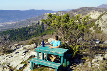 Fototapeta na wymiar A hiker who went on a mountain hike sits at a table in a picnic area and enjoys the view