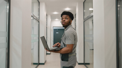 Fototapeta na wymiar African American installer in uniform stands in hallway and sets up security cameras using program on laptop. CCTV cameras installation in coworking office. Concept of surveillance system.