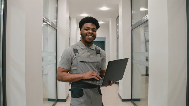 African CCTV cameras installer in uniform stands in corridor, smiles and looks at camera. He sets up security cameras in office using software on laptop. Concept of surveillance system.