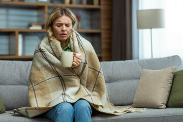 Obraz premium Tired senior woman sitting at home on the sofa covered with a warm blanket. Holds a cup of hot drink, medicine. He is sick with the flu, a cold. He feels pain, suffers, has no strength.