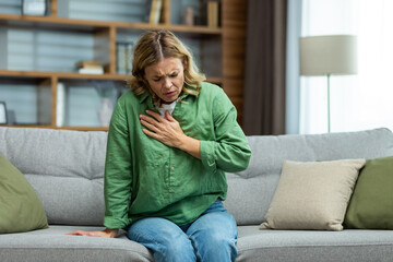 Senior beautiful woman sitting sick on sofa at home. Holds his chest. He has asthma, allergies, feels severe pain, tenderness, panic attack, infarct. - obrazy, fototapety, plakaty