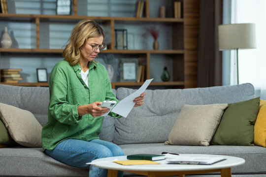 Senior beautiful woman in glasses sitting on sofa at home. Worriedly holding the phone and documents in his hands. Problem with accounts, credit, payment.