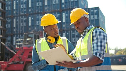 Two professional african engineers wearing safety uniform and hard hats working on laptop and holding a communication radio at cargo
