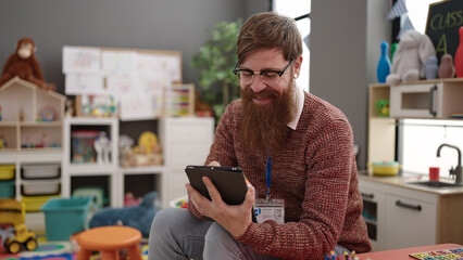 Young redhead man preschool teacher writing on touchpad sitting on chair at kindergarten