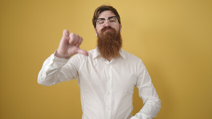 Young redhead man doing negative gesture with thumb down over isolated yellow background