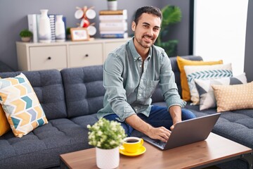 Young hispanic man using laptop drinking coffee at home