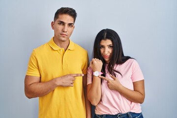 Young couple standing over isolated background in hurry pointing to watch time, impatience, looking at the camera with relaxed expression