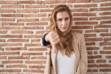 Beautiful blonde woman standing over bricks wall looking unhappy and angry showing rejection and negative with thumbs down gesture. bad expression.