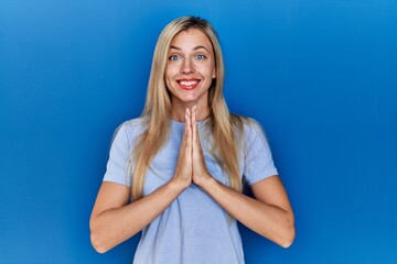 Beautiful blonde woman wearing casual t shirt over blue background praying with hands together asking for forgiveness smiling confident.